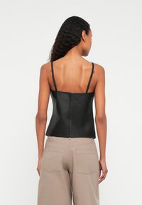 Pikkade lokkis juustega naine seisab seljaga, kandes musta nahast ilma varrukateta toppi ja beeže kõrge vöökohaga pükse.