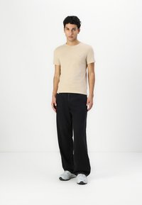 Camiseta de manga corta beige con un pequeño logo rojo, combinada con pantalones anchos negros y zapatillas grises. Diseño sencillo con una forma ajustada.