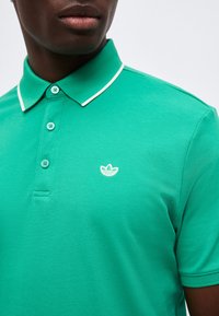 Άνδρας που φοράει πράσινο πόλο με άσπρη μπορντούρα στο γιακά και μικρό λευκό κεντητό λογότυπο στο αριστερό στήθος.