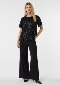 Czarna cekinowa bluzka z okrągłym dekoltem, zestawiona z szerokimi czarnymi spodniami i czarnymi butami na szpic; gładka faktura z błyszczącymi ozdobami.