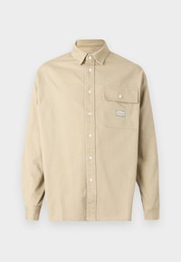 Maglietta beige a maniche lunghe con un colletto appuntito e una tasca con patta abbottonata che presenta una piccola toppa decorativa.