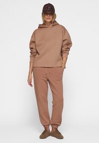 Brun sweatshirt med hætte og snørelukning, matchende tapered sweatpants og brun teksturerede sneakers. Kasketten har leopardmønster.