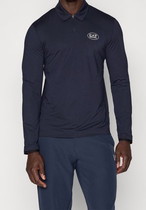 Hombre que lleva una camiseta polo azul marino de manga larga con cremallera y logo EA7 en el pecho y pantalones azul marino a juego.