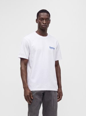 Un mannequin homme se tient devant un fond de studio gris clair, vêtu d’un T-shirt blanc Dickies et d’un pantalon gris.