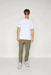 Witte t-shirt met korte mouwen en een borstzak, voorzien van een lichtblauwe accent, gecombineerd met olijfgroene tapered broek en grijze sneakers.