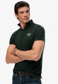 Donkergroen poloshirt met een kraag met knopen en een geborduurd logo op de borst, gecombineerd met donkere denim jeans en een bruine riem.