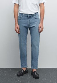 Svetle modré džíny s rovnými nohavicemi, pěti kapsami a zapínáním na knoflík a zip, nošené s bílou košilí a černými botami.