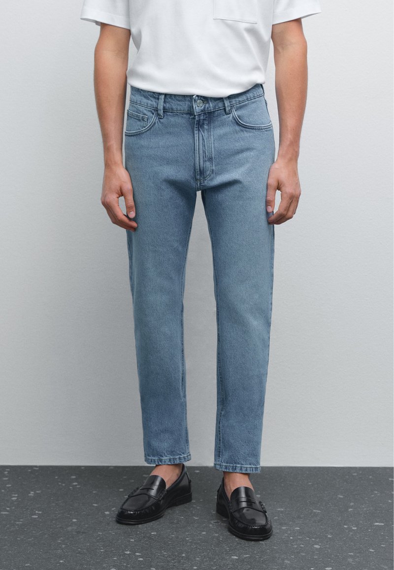 Svetle modré džíny s rovnými nohavicemi, pěti kapsami a zapínáním na knoflík a zip, nošené s bílou košilí a černými botami.