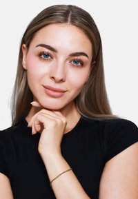 Une femme aux cheveux raides châtain clair et aux yeux bleus repose son menton sur sa main, portant un tee-shirt noir et un bracelet en or fin.