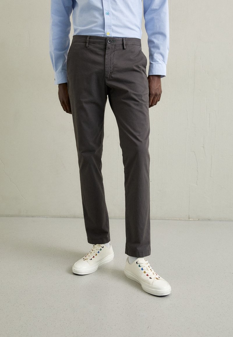 Homme portant un pantalon gris taille fine, une chemise à boutons bleu clair, des baskets blanches avec des œillets multicolores, debout sur un sol gris clair.