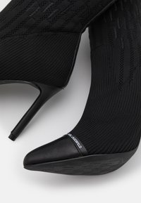 Chaussures noires à talons hauts avec un dessus en tissu tricoté, un bout pointu et des accents en cuir. Comprend une étiquette logo au niveau de la cheville.