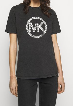 Personne portant un T-shirt noir à manches courtes avec un cercle en pointillés et le logo "MK" sur le devant, associé à un pantalon noir.