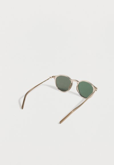 Στρογγυλά γυαλιά ηλίου με χρυσά μεταλλικά πλαίσια και σκουροπράσινους φακούς που ακουμπούν σε λευκή επιφάνεια.