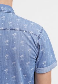 Camicia in denim azzurro chiaro con un motivo ripetuto di palme bianche e piccoli punti bianchi, maniche corte arrotolate e colletto a punta.