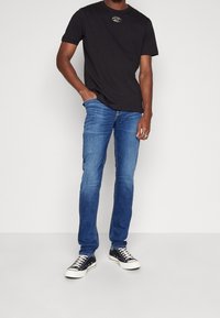 Camiseta de algodón negra con un pequeño logo plateado, combinada con unos vaqueros de mezclilla azules. El modelo lleva zapatillas de lona negras con suelas blancas.