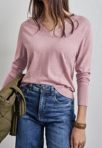 Rosa lättviktströja med V-ringning och ribbade ärmar, tillsammans med mörkblå jeans och en olivgrön pufferjacka hängande över armen.