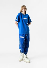 Camiseta de algodón azul con cuello redondo y mangas cortas, combinada con pantalones de chándal azules que presentan un diseño bordado y acentos blancos.