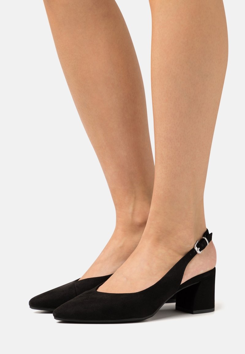 Jambes portant des chaussures à talons blocs en daim noir avec une boucle en argent sur fond clair, montrant une vue de côté.