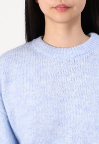 Pull bleu clair en maille avec un col rond côtelé ; texture douce, coupe légèrement ample et motif de tricot visible.
