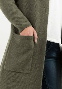 Oliven grøn strik cardigan med ribbet tekstur, lange ærmer og en stor lomme på venstre side. Kombineret med en hvid top og blå jeans.