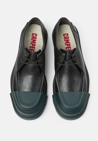 Schwarze Lederschuhe mit strukturiertem Finish, ausgestattet mit einer grünen Gummikappe, Schnürdesign und rotem Logo auf der Innensohle.