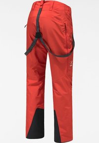 Pantalon d'extérieur rouge vif avec des renforts noirs à l'intérieur des chevilles et des sangles grises de harnais d'escalade attachées, vu de dos.