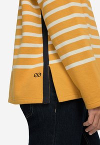 Sweatshirt giallo a righe con strisce bianche, caratterizzato da uno spacco laterale e un accento blu navy. Realizzato in tessuto morbido, abbinato a jeans scuri.