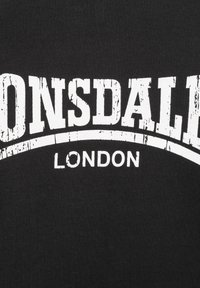Čierne tričko s odvážnym bielym textom "LONSDALE" nad "LONDON", so zničenou textúrou a zakriveným dizajnom pod nápisom.