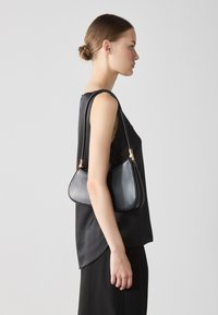 Black leather handbag with a curved shape, dual shoulder straps, and gold-tone hardware details, worn on the shoulder against a neutral background.