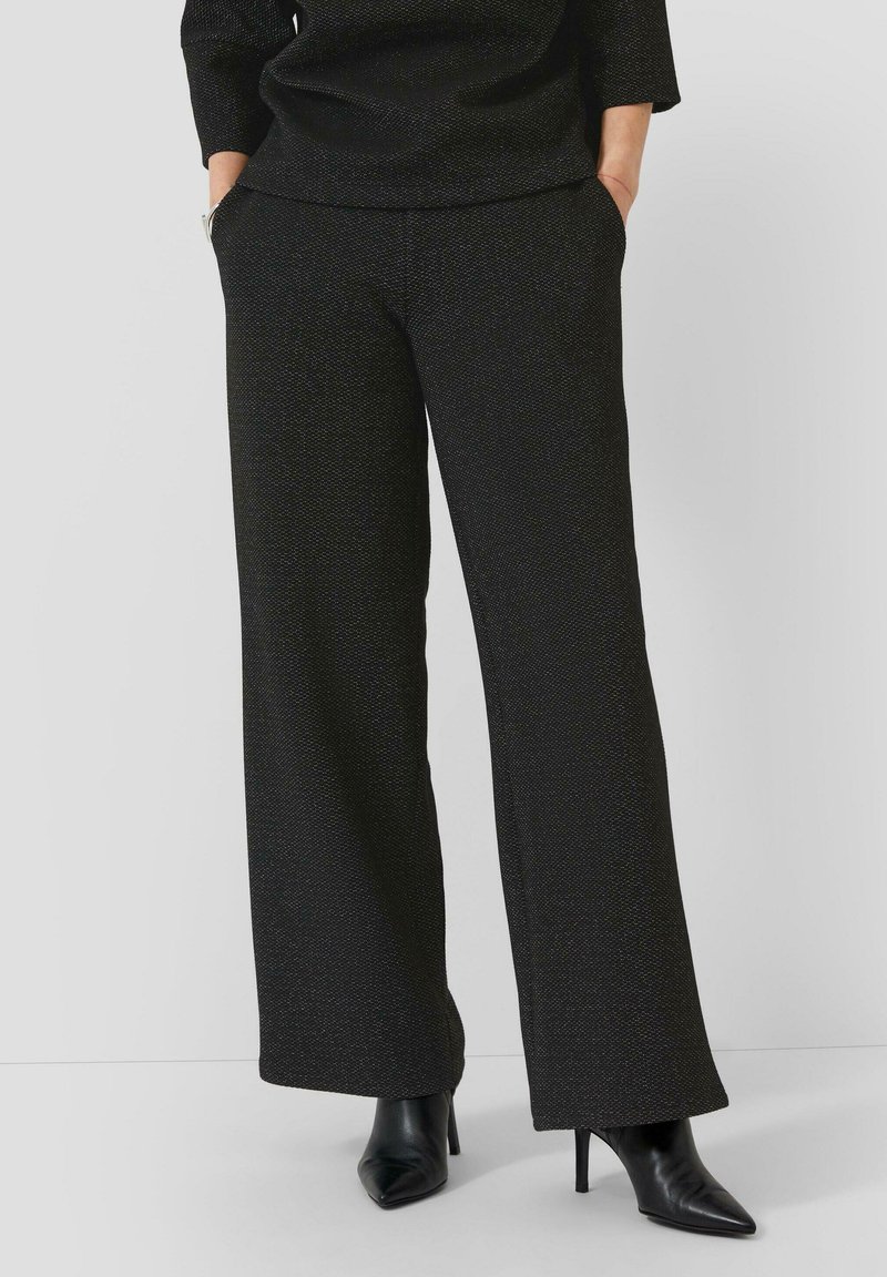 Femme portant un pantalon large texturé noir avec les mains dans les poches et des bottes à talons aiguilles noires sur fond blanc.
