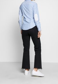 Camicia azzurra chiara con toppe ai gomiti bianche e texturizzate, abbinata a jeans neri a palazzo con orli sfilacciati e sneakers bianche.