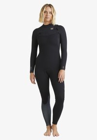 Frau steht barfuß in einem enganliegenden schwarzen Neoprenanzug mit langen Ärmeln und einem Reißverschluss auf der Vorderseite, vor einem schlichten weißen Hintergrund.