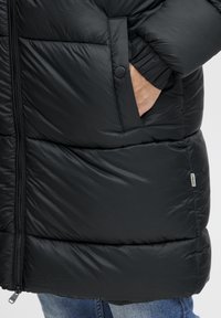 Chaqueta puffer negra con un acabado texturizado, cierre de cremallera, bolsillos laterales y puños elásticos, con una sutil etiqueta de la marca en el dobladillo inferior.