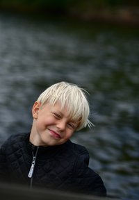 Child with short, spiky blonde hair, wearing a black quilted jacket with a zip. Background shows blurred water. Child is smiling playfully.