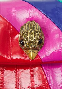 Värikäs käsilaukku, jossa on punaisia, vaaleanpunaisia ja sinisiä osioita kiiltävällä pinnalla. Huomattava kultainen lintukuvioinen koriste, jossa mustat silmät.