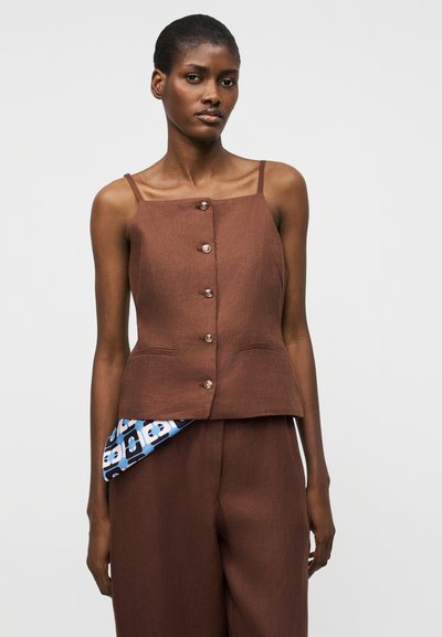 Γυναίκα που φοράει καφέ αμάνικο τοπ με κουμπιά και λεπτές τιράντες, καθώς και παντελόνι μέσης σε αντίστοιχο χρώμα, με μπλε ύφασμα με σχέδιο που ξεπροβάλλει από την τσέπη.