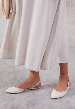 Ankel-længde lyse nederdel med hvide spidse slingback-flade sko prydet med guld, der viser tatoveringer på begge ankler.