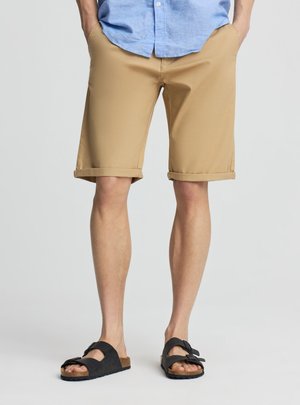 Uomo che indossa pantaloncini kaki risvoltati, camicia azzurro chiaro con bottoni e sandali neri con punta aperta, in piedi con le mani nelle tasche.