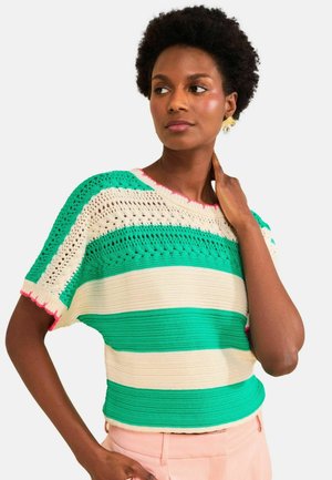 Woman with short curly hair wearing green and cream striped knit top with decorative holes and pink-trimmed sleeves, posing with hand on neck.