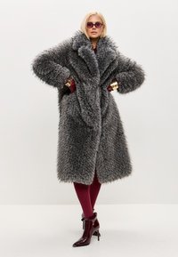 Falsk pelsfrakke i grå med bred krave og voluminøse ærmer. Båret over bordeaux strømpebukser og skinnende ankelstøvler, der skaber et lagdelt look.