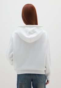 Sudadera blanca con una textura suave, corte amplio y capucha con cordón. Combinada con jeans azules, mostrando un estilo casual y relajado.