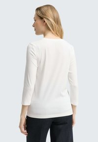 Weißes Langarmoberteil mit rundem Halsausschnitt und glattem, leichtem Stoff; von hinten gezeigt, kombiniert mit dunklen Jeanshosen.