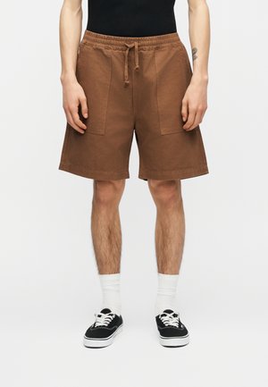 Mand iført brune knælange shorts med forlommer, hvide sokker, sorte sneakers og en sort skjorte mod en ensfarvet baggrund.