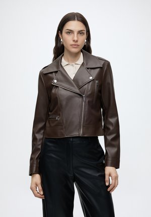 Mujer con chaqueta de cuero marrón oscuro estilo motorista sobre camisa beige y pantalones negros, de pie frente a un fondo claro y sencillo.