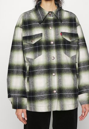 Personne portant une veste à carreaux surdimensionnée verte, noire et blanche avec de gros boutons et deux poches poitrine, debout devant un fond uni.