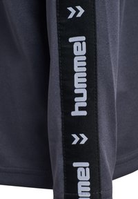 Mørkegråt stof med sort lodret stribe, der viser hvidt "hummel"-logo og chevron-symboler langs ærmet eller buksebenet.