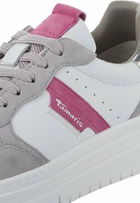 Baskets blanches avec des accents en daim gris et une bande rose. Présente des lacets texturés, des trous d'aération à pois, et un logo sur le côté. Semelle lisse.