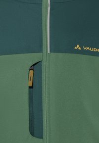 Giacca verde con zip caratterizzata da un design bicolore con pannelli verde scuro e verde chiaro, una tasca con zip e un logo giallo. Tessuto testurizzato.