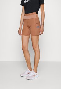 Shorts ajustés marron avec une taille haute et une poche latérale, arborant le logo bleu de Nike Trail. Portés avec un T-shirt noir et des baskets rose pâle.