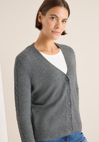 Cardigan grigio a maglia con scollatura a V e chiusura a bottoni. Presenta maniche a coste con motivo a treccia e orlo a coste. Materiale morbido e caldo.